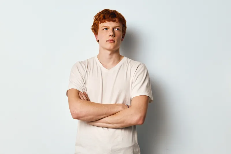 Young man with red hair and freckles wearing white t shirt standing with arms crossed against light background, looking away with confident expression in studio.