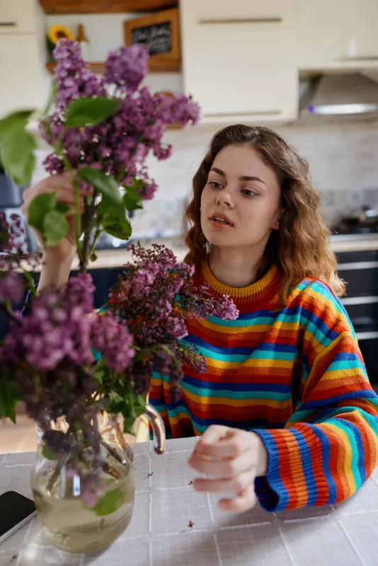 A woman in a colorful striped sweater arranges purple flowers in a kitchen vase, bright natural light fills a cozy home, illustrating calm domestic life and simple flower care.