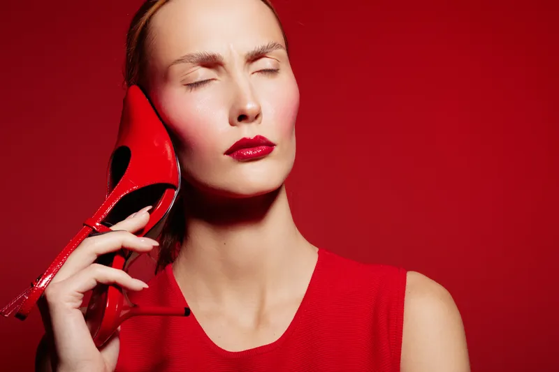 Woman in red dress holds red high heel shoe to her face with closed eyes, expressing discomfort or pain. Red background and matching colors create a bold, dramatic mood.