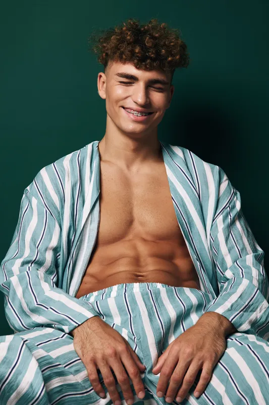 Young muscular man with curly hair and braces, smiling dressed in striped relaxed pajamas posing confidently against green studio background for lifestyle and wellness.