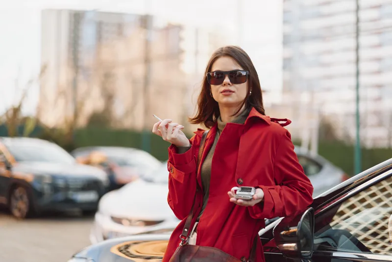 A confident young woman in a red coat and sunglasses smokes a cigarette while standing next to a car in an urban environment during daytime.