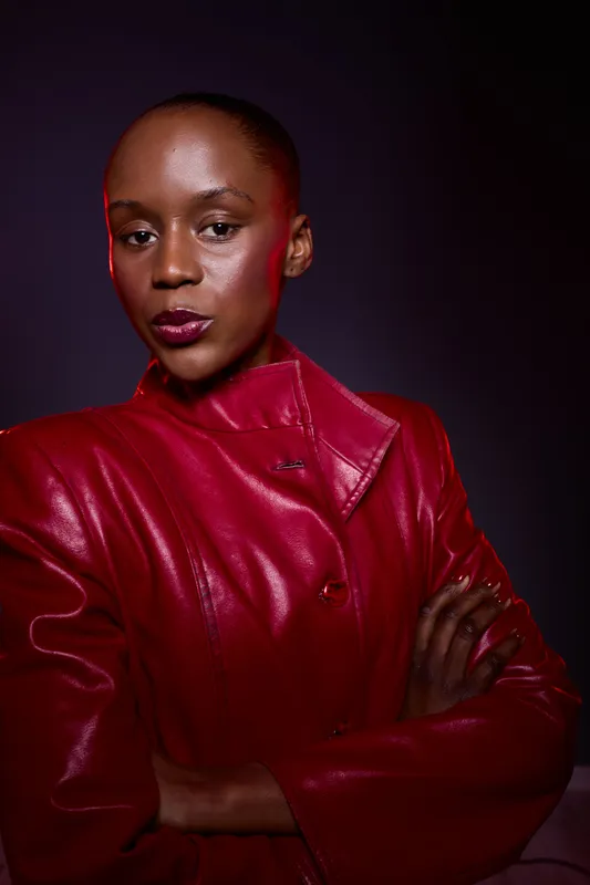 Confident dark skinned African woman in red leather jacket poses with crossed arms against dark background. Studio portrait emphasizing fashion, strength, and elegance.