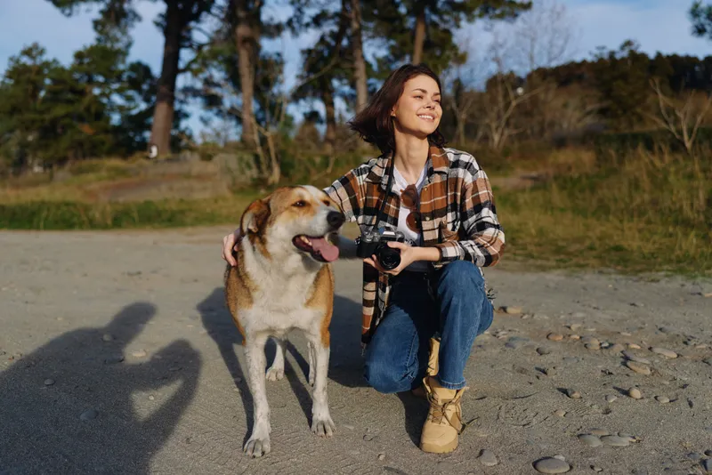 Woman with dog outdoors kneels on a gravel path, smiling as she holds the leash, enjoying a sunny day in a park with trees, open space, and a gentle breeze.