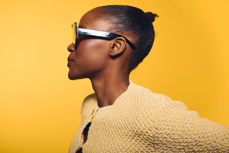 Profile of a confident African woman with dark skin wearing large sunglasses and a textured yellow sweater. Bright yellow background emphasizes fashion and modern style concept.