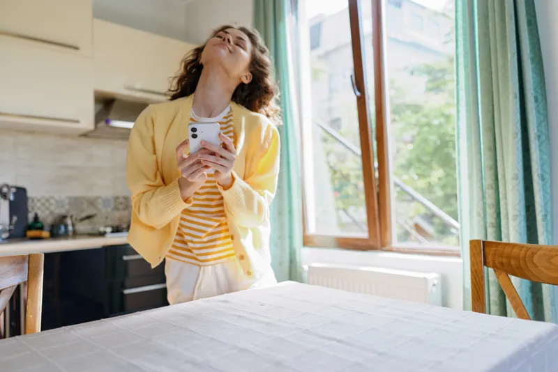 A young woman in a yellow cardigan basks in warm natural light in a bright kitchen, smiling softly as she takes a casual selfie, showing a relaxed, joyful mood.