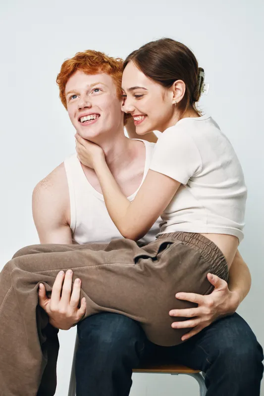 Young couple smiling and embracing in casual clothing. Woman sitting on man’s lap with happy expressions for love, friendship and relationship. Indoor studio with light background.