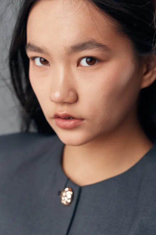 Portrait of young woman with serious expression and natural makeup wearing dark clothing against neutral background. Closeup face with soft light highlighting skin texture and eyes.