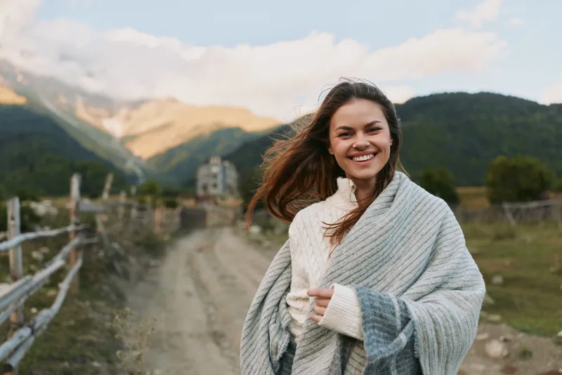 Woman enjoying outdoor lifestyle in mountains, smiling, wrapped in scarf and sweater, bright scenic countryside, fresh air and natural light capture