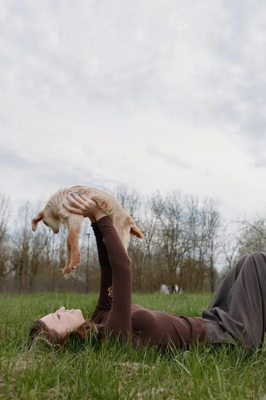Woman lies on grass outdoors, tossing a young goat into the air, smiling softly, casual outfit, carefree moment, natural colors, rural park vibe, sunlight on skin, fresh air, warm mood