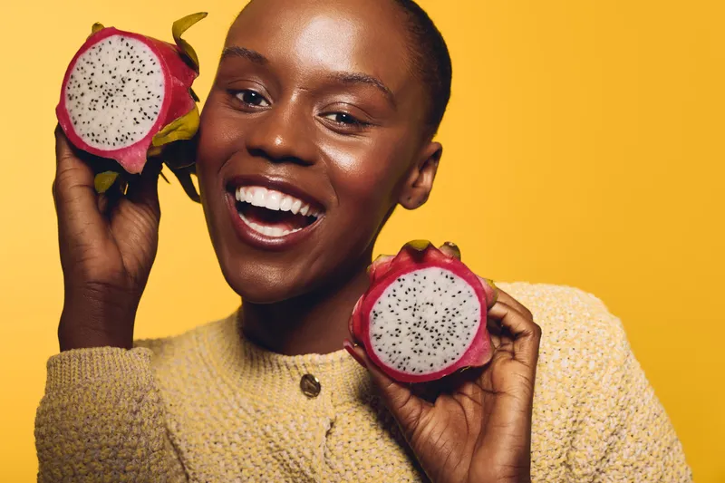 Smiling black African woman with dark skin holding two halves of a dragon fruit near her face, wearing a yellow sweater on a bright yellow background. Concept of healthy lifestyle and fresh fruit.