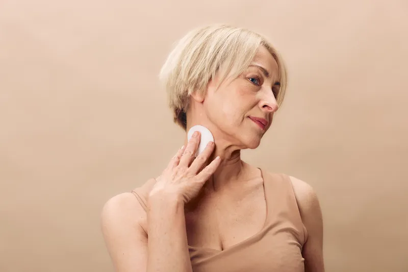 Mature woman with short blonde hair gently applying a round facial sponge on her neck. Neutral beige background and soft natural lighting create a calm skincare and self-care concept.