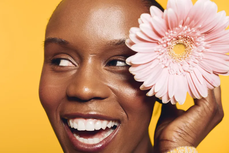 Close-up of smiling black woman with dark skin holding pink flower near face on yellow background. Concept of beauty, skincare, happiness, and natural cosmetics with floral element.
