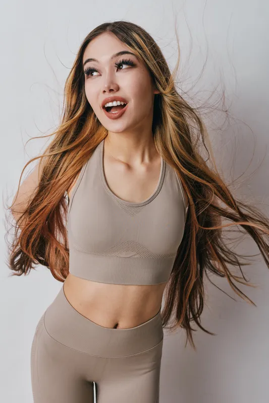 Young woman with long hair wearing beige sportswear posing indoors with happy expression. Fitness, health, casual outfit and beauty portrait with natural makeup and flowing hair in studio.