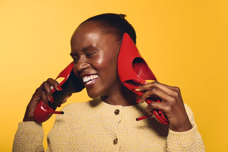 Happy African woman with dark skin laughs holding red high heels near face on yellow background. Concept of joy, fashion, lifestyle, and playful mood in studio portrait.