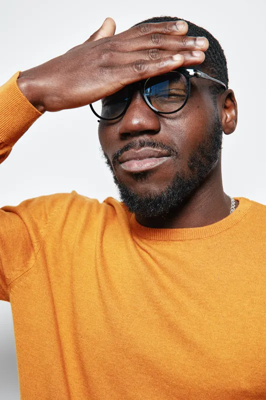 African man with dark skin wearing glasses and orange sweater covering forehead with hand, posing with closed eyes on white background. Portrait, casual style and thoughtful expression.