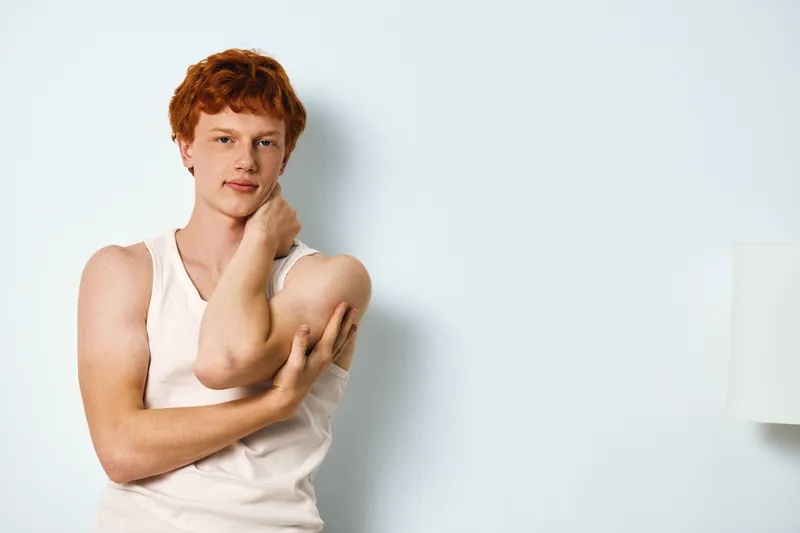 Young man with red hair wearing a white tank top poses with hand on neck against a plain light background in casual style and confident expression.