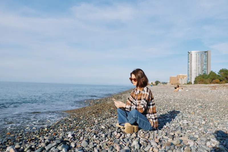 Woman sitting on a pebble beach with a plaid shirt, watching the sea and distant buildings under a clear sky. Casual moment of reflection, sea breeze, and calm coastal scenery.