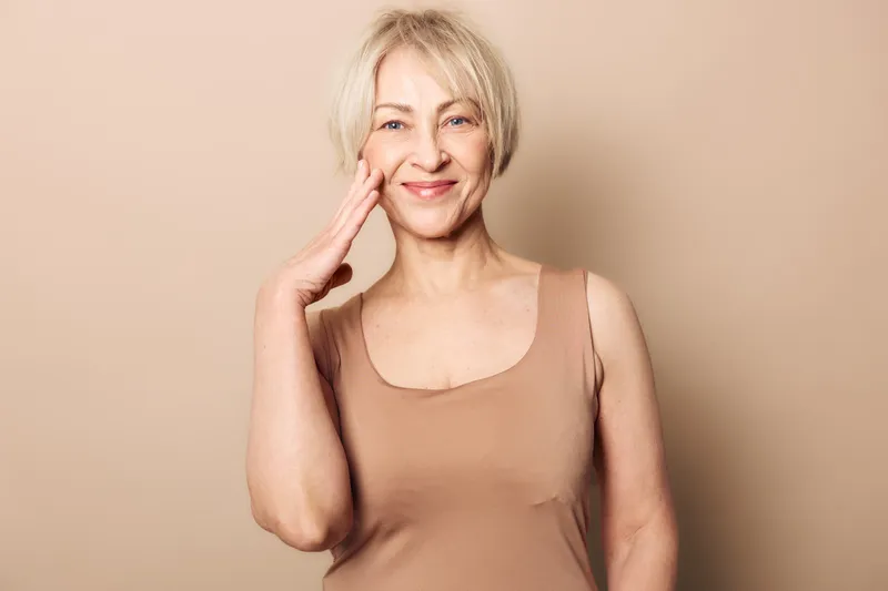 Smiling mature woman with short blonde hair touches her face gently against beige background. Concept of natural beauty, aging gracefully, skincare routine, confidence and wellness.