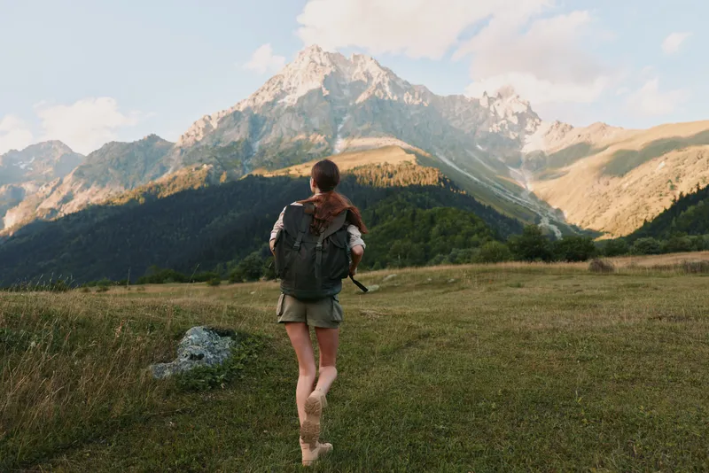 A young woman with a backpack walks across a grassy meadow toward distant rugged mountains, embodying adventure and freedom in a vast natural landscape under clear skies