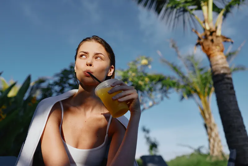 Cheerful woman outdoors enjoying a cold juice under bright sun, relaxed posture, palm trees in background, warm breeze, vibrant colors, carefree lifestyle moment today