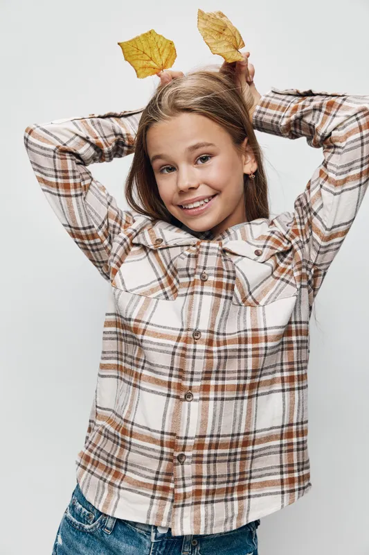 Smiling girl with brown hair holding autumn leaves behind her head wearing plaid shirt and jeans, posing on white background for fall season, happiness and childhood fun outdoors.