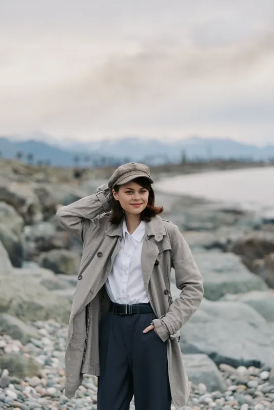 Young woman in knitted coat and cap stands on rocky beach near calm sea with mountains in background. Casual fashion and outdoor lifestyle concept with soft natural light.