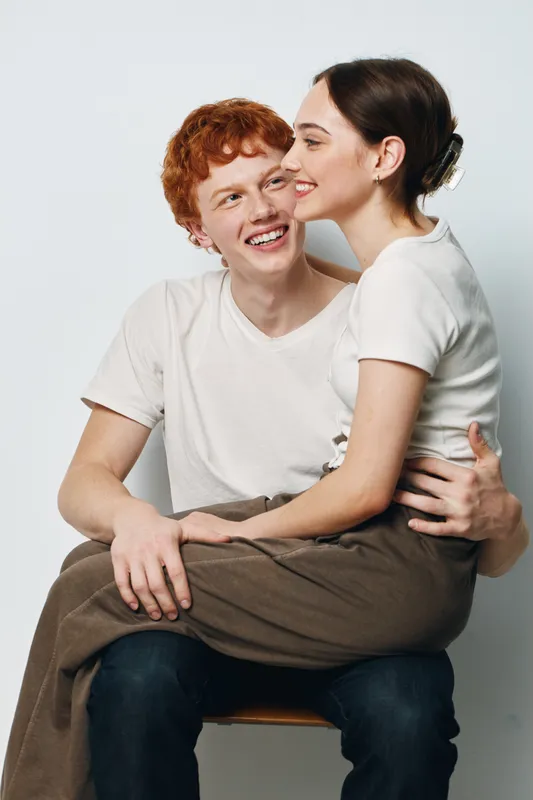 Couple sitting closely laughing with casual clothes white background studio. Young man and woman smiling happy affectionate in relaxed pose with love and friendship.