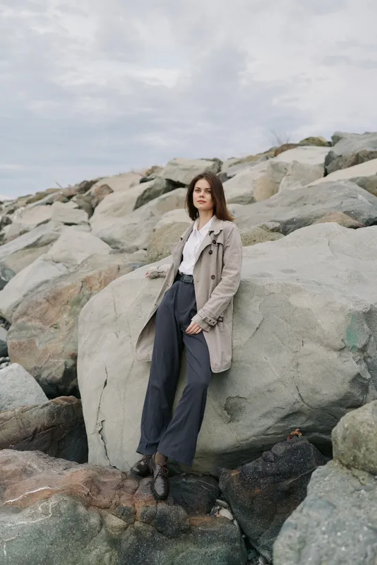 Young woman in coat and white shirt stands on rocky shore under cloudy sky. Casual fashion portrait with natural background. Concept of style, confidence, and outdoor mood.