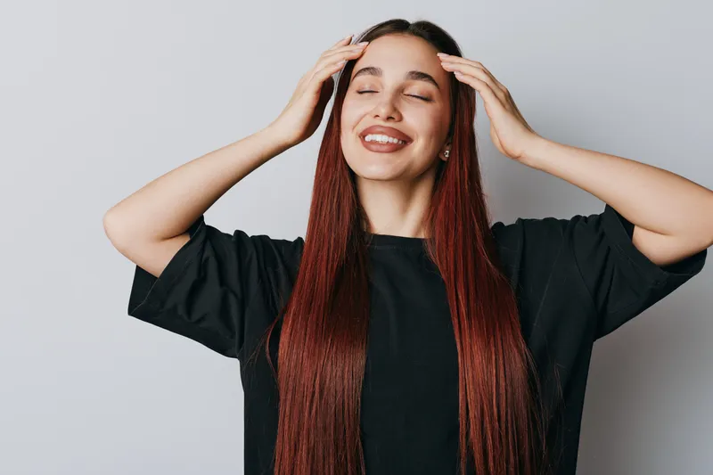 Woman with long red hair and closed eyes smiling gently with hands touching head wearing black shirt on light background. Relaxed female portrait with natural makeup and earrings indoors.