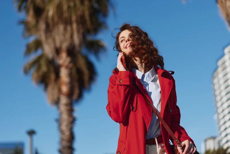 Young woman with curly hair wearing a red coat smiles while talking on the phone outdoors on a sunny day with palm trees in the background.
