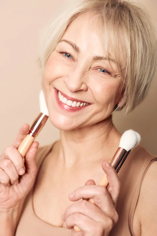 Smiling mature woman with short blonde hair holds two makeup brushes near face, showing natural beauty and skincare concept with soft lighting and warm beige background.