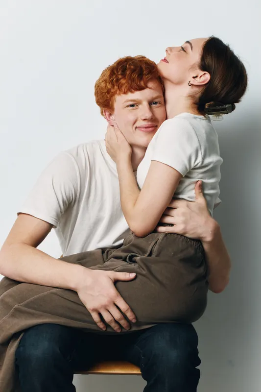 Couple, young man and woman hugging and smiling in casual clothes sitting on chair with white background for love and relationship portrait.