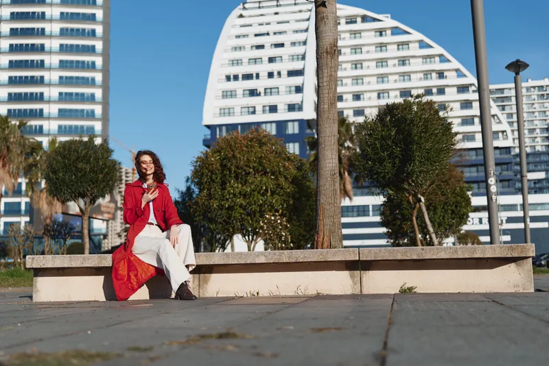 Cheerful woman outdoors wearing a red coat and dress, seated on a low wall in a sunny city plaza, with modern architecture, palm trees, and blue sky in the background