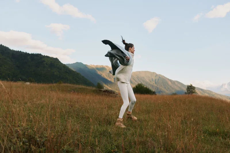 Young woman jumping in a grassy field with mountains in the background. Outdoor lifestyle concept with casual clothing and natural light during daytime.