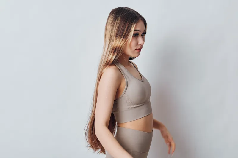 Young woman with long hair wearing beige sportswear stands against plain background in profile. Fitness, wellness and casual active lifestyle concept with natural light and minimal style.