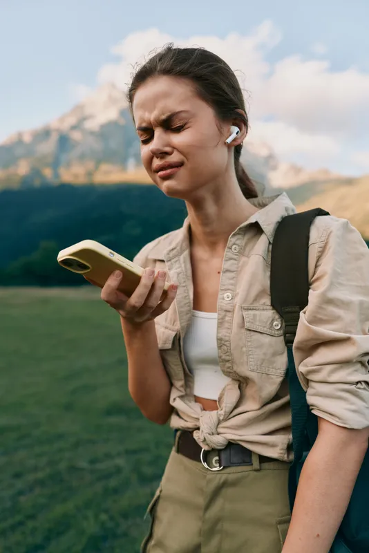 Smiling woman outdoors checking smartphone during a hike. Casual outfit and backpack against a mountainous landscape. The scene conveys lifestyle, exploration, and wellness.