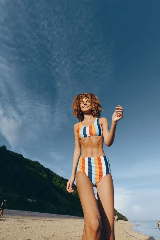 A smiling woman in a colorful striped bikini stands on a sandy beach with green hills and a bright blue sky in the background. Concept for summer, vacation, and outdoor lifestyle.