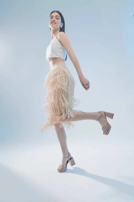 Young woman in a stylish white top and fringed skirt poses gracefully on one leg against a light blue background. Concept for woman beauty fashion and elegance.