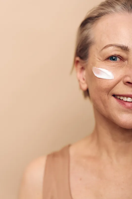 Mature woman with smooth skin smiling applying moisturizing cream on cheek against beige background. Concept of skincare routine, anti aging, self care and natural beauty for healthy skin.