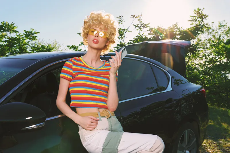 Smiling woman with curly hair leaning against a dark car, wearing a striped colorful top. Outdoor scene with warm sun, retro film color tone, and a casual lifestyle vibe.