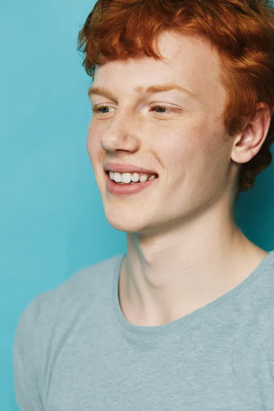 Portrait of a smiling redhead teenage boy with freckles wearing a gray shirt on a blue background. Casual youth with natural hair looking aside with happiness and confidence.