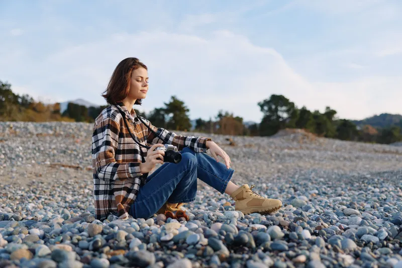Woman sitting on a pebble beach with a camera, wearing a plaid shirt and jeans, a tranquil outdoor scene suitable for photography, travel and nature lifestyle projects