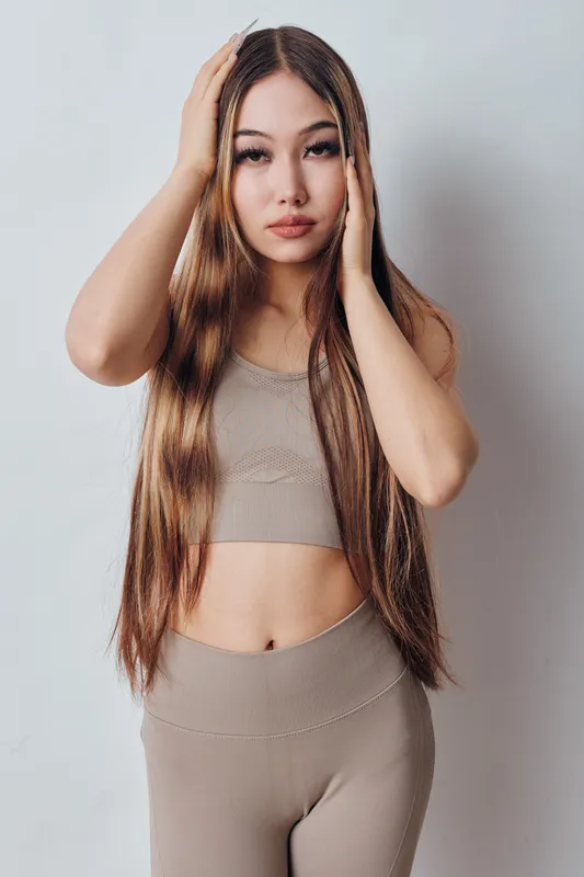 Woman with long hair wearing beige crop top and leggings posing with hands near head against light plain background in studio with natural makeup and serious expression.
