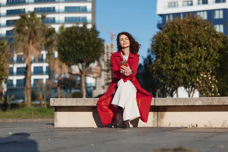 A stylish young woman sits on a concrete bench in an urban park, wearing a red coat and white dress, posing with confidence in daylight.