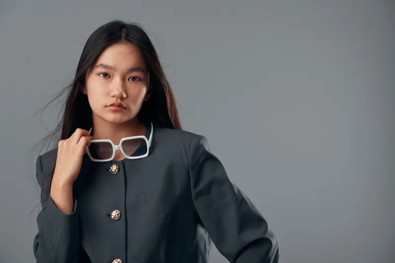 Woman with long dark hair wearing blazer holds white sunglasses by collar posing against gray background in studio for fashion portrait and style concept with serious expression.
