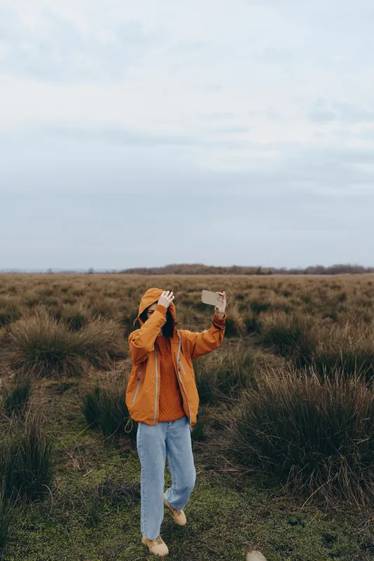 Woman lifestyle outdoors, standing and walking in a grassy field under a cloudy sky, orange jacket and jeans, holding paper and phone, calm nature moment, gentle daylight wind