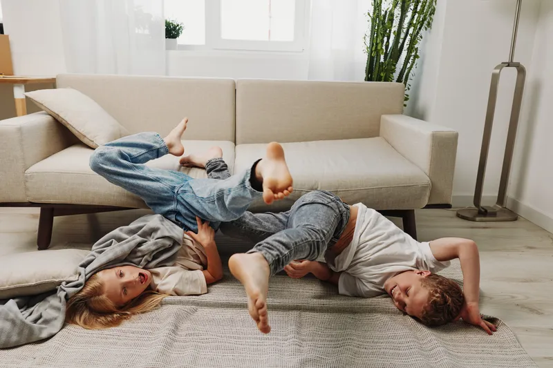 Two playful children in casual clothes tumble and laugh on a soft rug in a bright living room with modern decor and natural light.