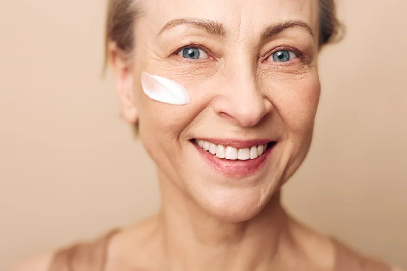Close-up of smiling mature woman with natural skin applying cream on cheek. Concept of skincare, anti aging, self care, beauty routine, and healthy glowing skin.
