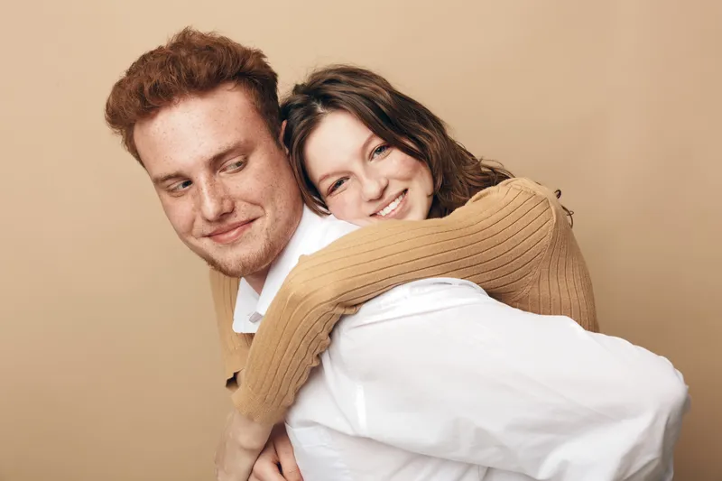 Smiling young woman in brown sweater hugs happy man with red hair and white shirt from behind. Warm beige background, close-up portrait of affectionate couple showing love and friendship.