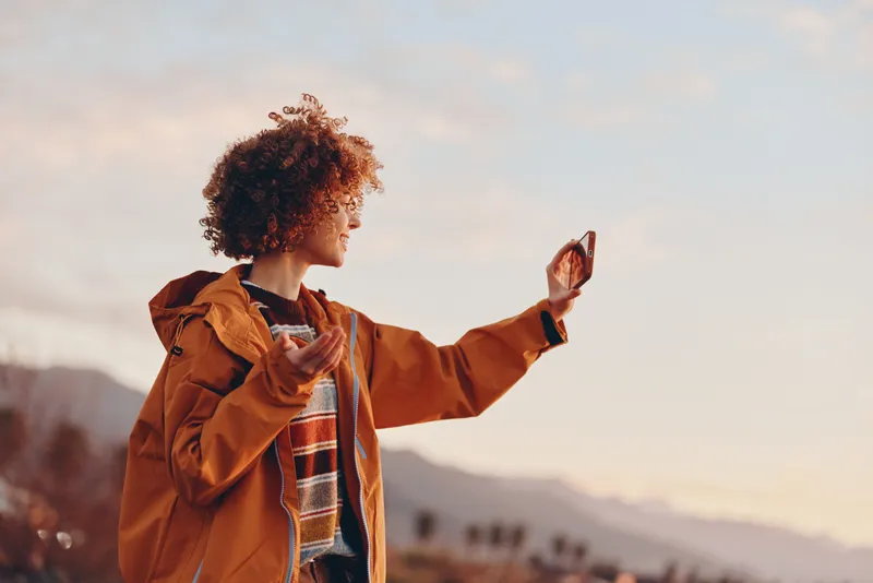 Woman lifestyle outdoors, curly hair orange jacket, holding smartphone for selfie at sunset, warm light, mountains background, relaxed confident travel mood, soft breeze, natural colors